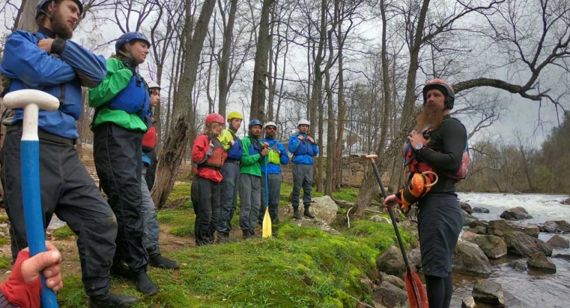 A group of people wearing lifejackets stand on the bank of a river, listening to an instructor. 